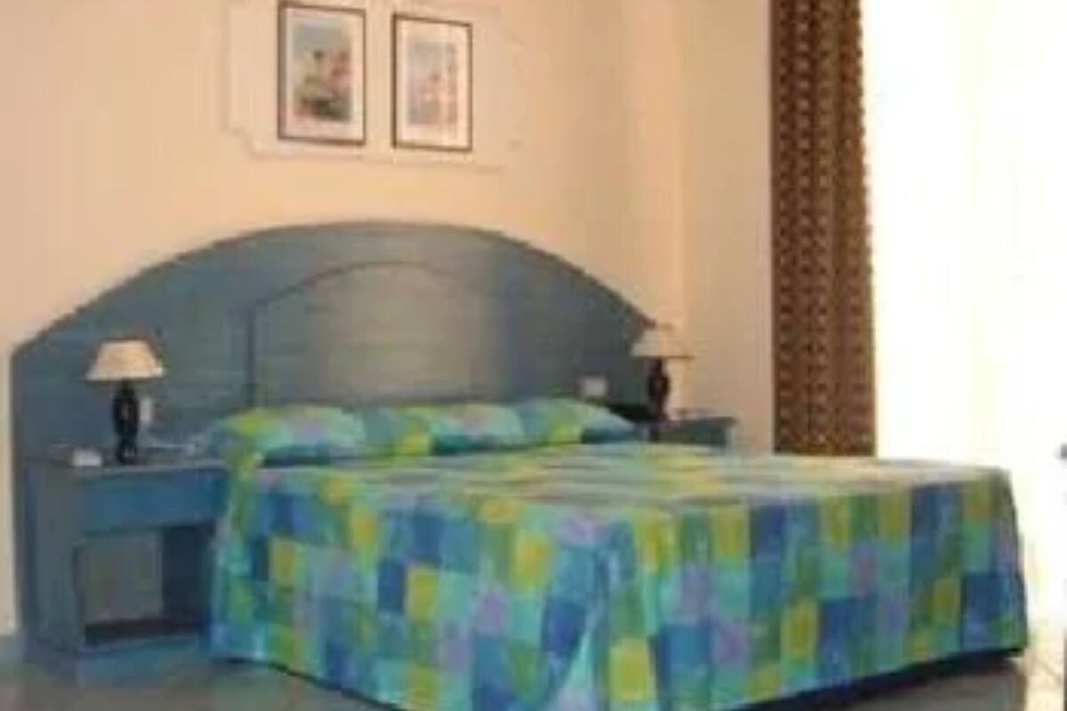 Ein Schlafzimmer mit blauem Kopfteil, einem Bett mit bunter Decke, zwei Nachttischen mit Lampen und zwei gerahmten Bildern an der Wand.