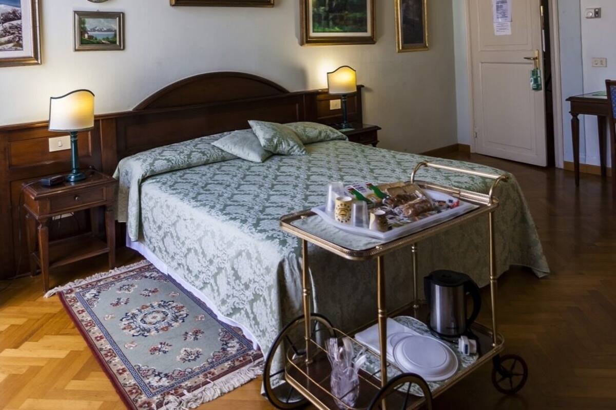 A hotel room with a wooden bed, a nightstand with a lamp, a bedside table, a tray table with breakfast items, a decorative rug, and framed pictures on the wall.