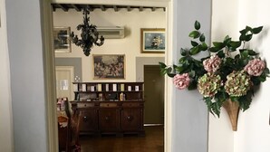 A hallway with a wooden floor leading to a dining area with a wooden table and chairs. There is a decorative plant on the wall and a chandelier above the dining table.