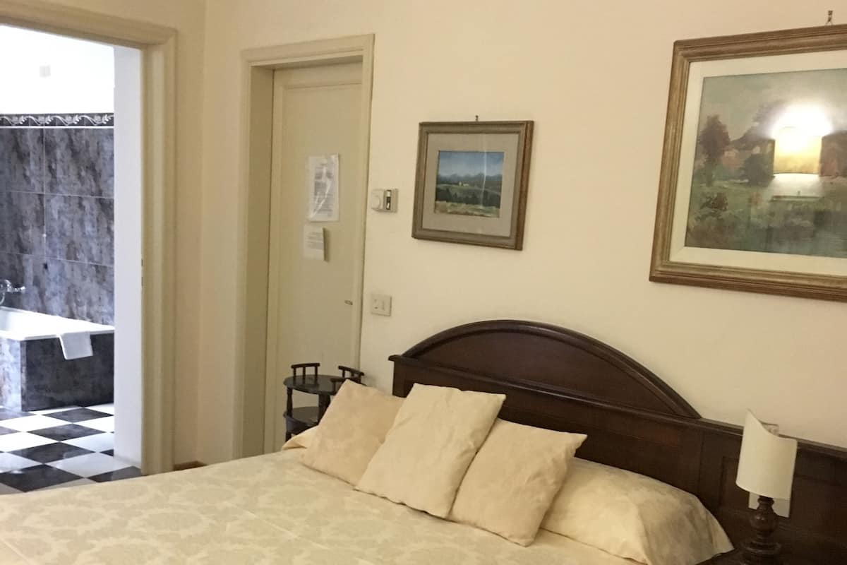 A bedroom with a large bed, a nightstand, a bedside lamp, a framed picture on the wall, and a door leading to another room.