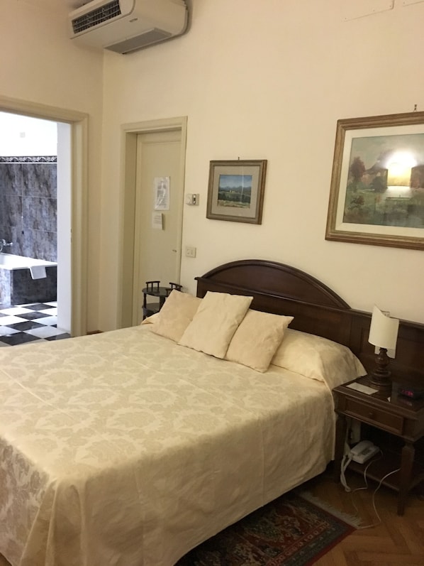 Un dormitorio con una cama grande, una mesita de noche, una lámpara de noche, un cuadro enmarcado en la pared y una puerta que da a otra habitación.