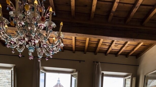 A room with a wooden ceiling, a chandelier, two beds, a chair, and large windows with a view of buildings.