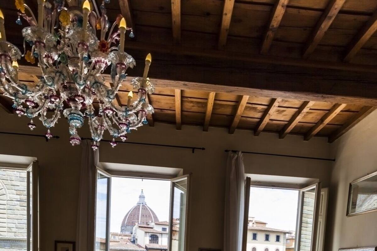 A room with a wooden ceiling, a chandelier, two beds, a chair, and large windows with a view of buildings.