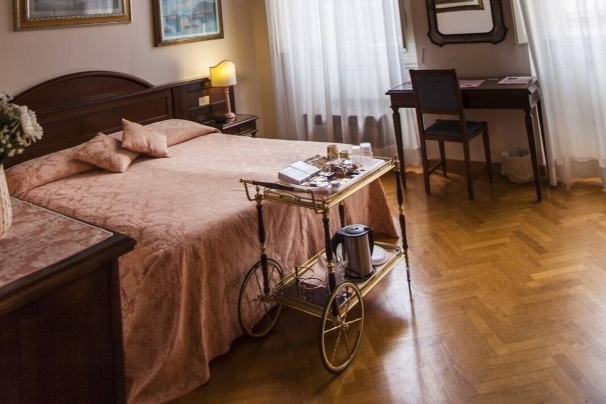 A room with a wooden ceiling, a bed with a pink bedspread, a desk with a chair, and a cart with items.