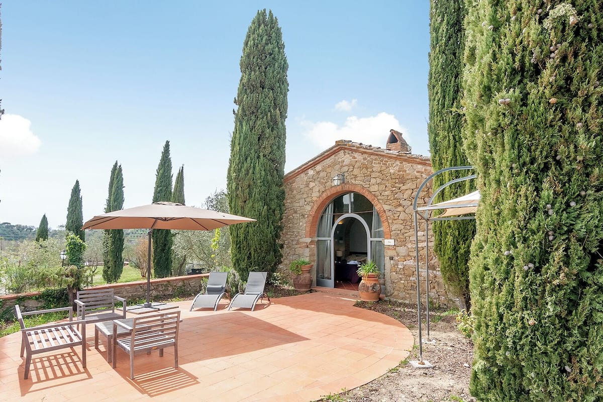A stone building with an arched entrance, surrounded by tall cypress trees and a patio area with outdoor seating and umbrellas.