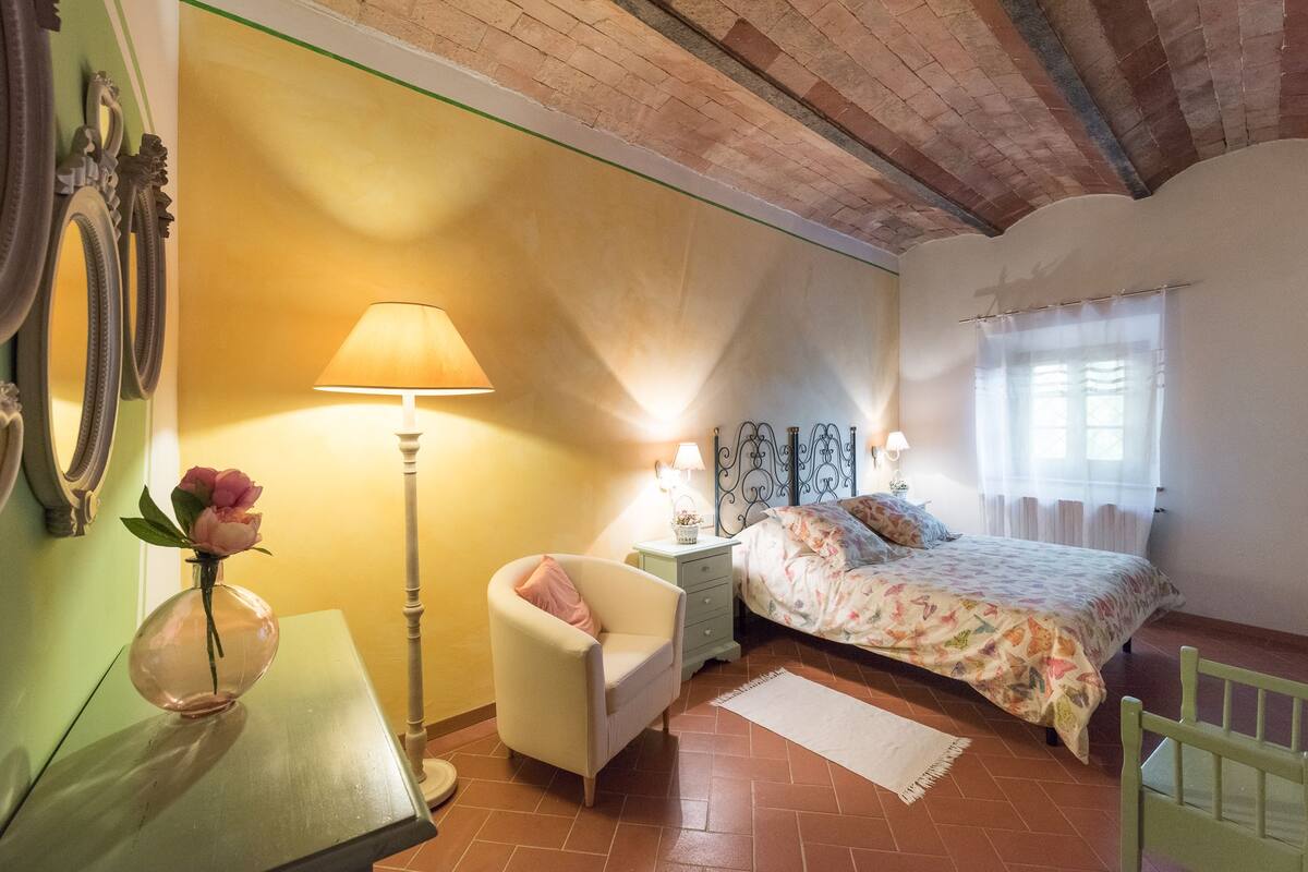A bedroom with a bed, a chair, a nightstand, a lamp, and a vase with a flower.