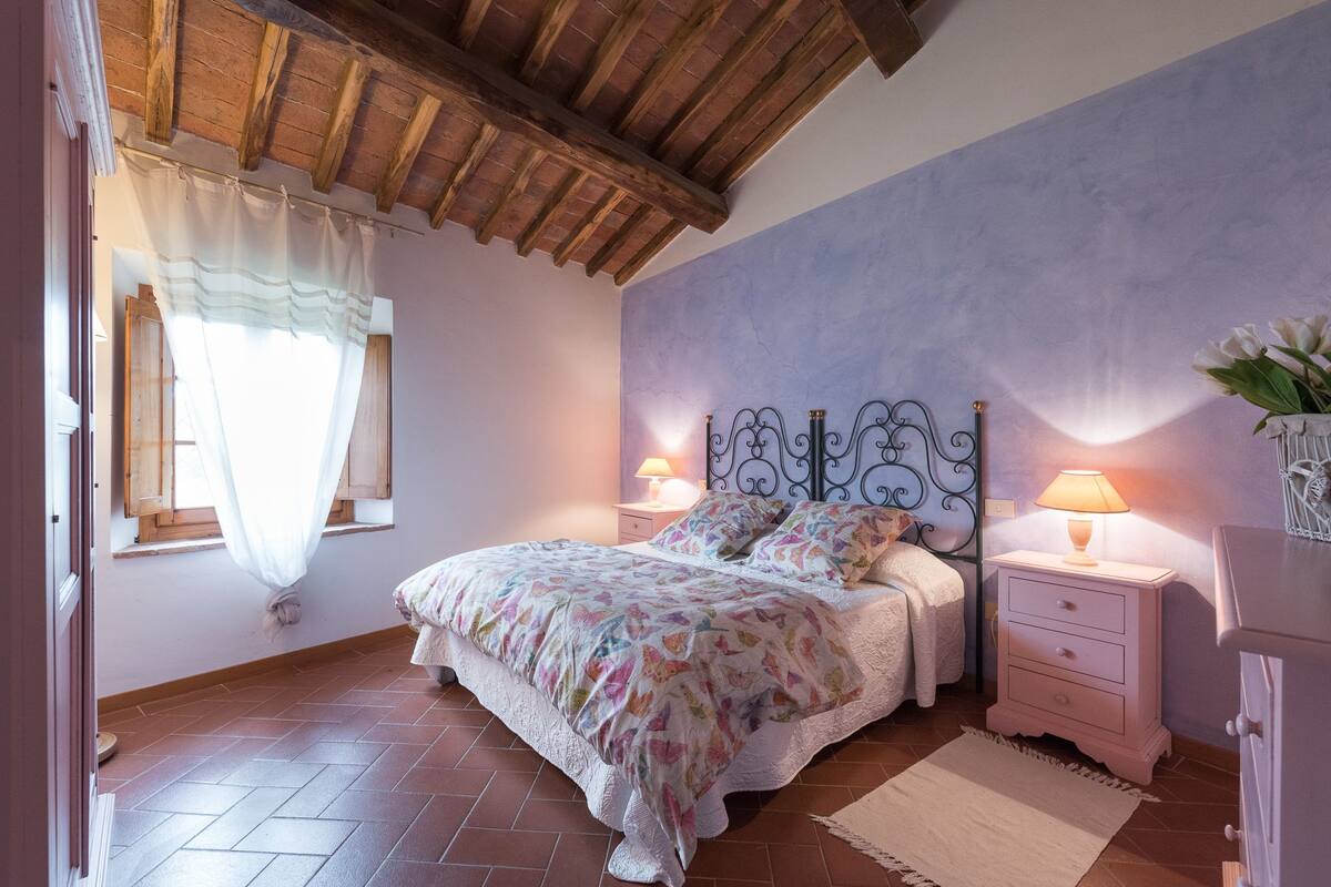A bedroom with a bed, two bedside tables with lamps, a window with curtains, and a wooden ceiling.
