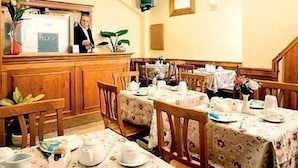 A dining area with wooden tables and chairs, a counter with a person behind it, and a small plant on the counter. - Hotel Kriss (Rome)