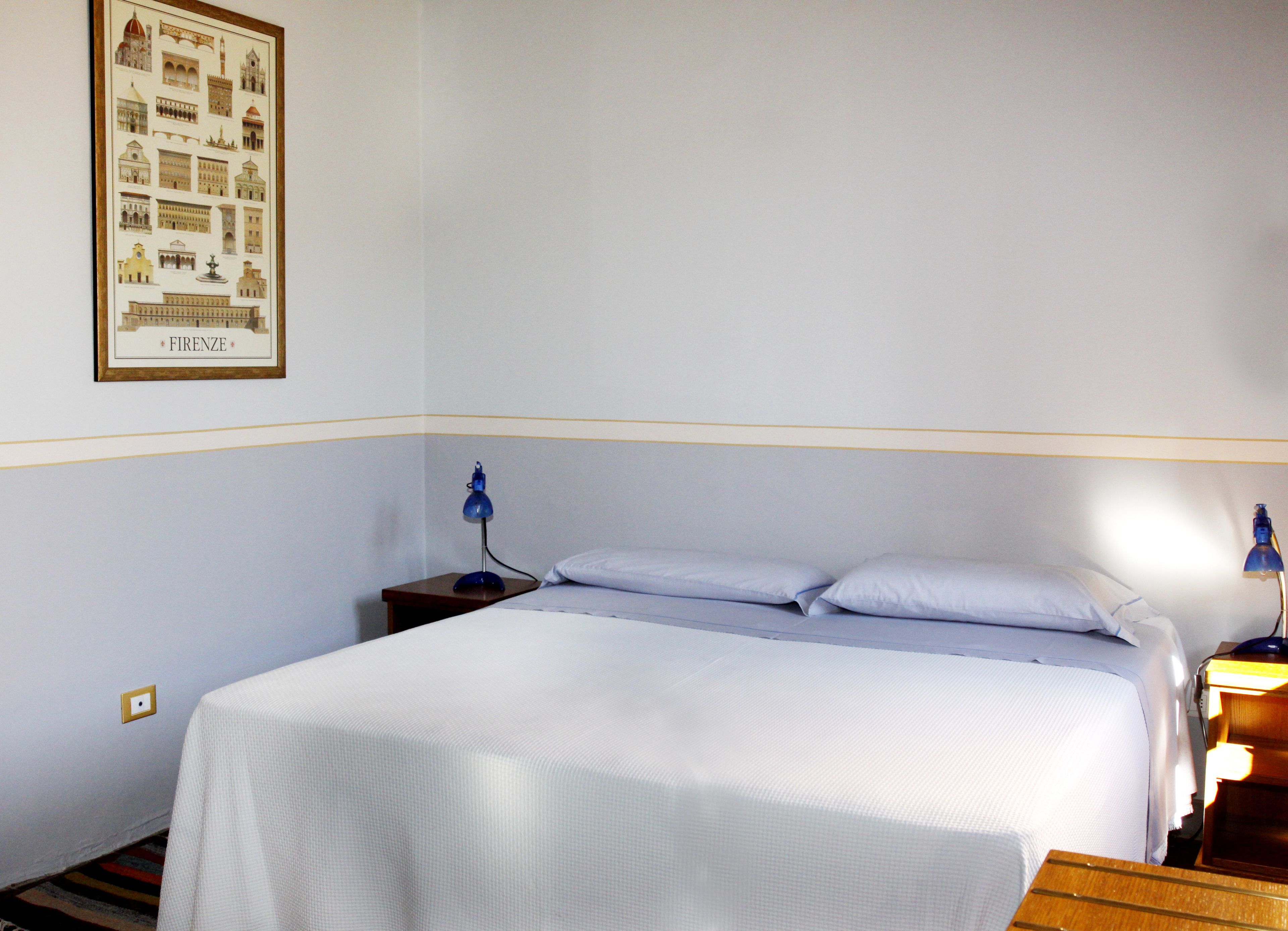 A hotel room with a bed, two bedside tables, a nightstand, a framed picture of Italian architecture, and a lamp on each table.