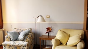 A living room with a floral-patterned armchair, a plain armchair, a floor lamp, and a small table with a vase of flowers. - Gianna's B&B (Florence)