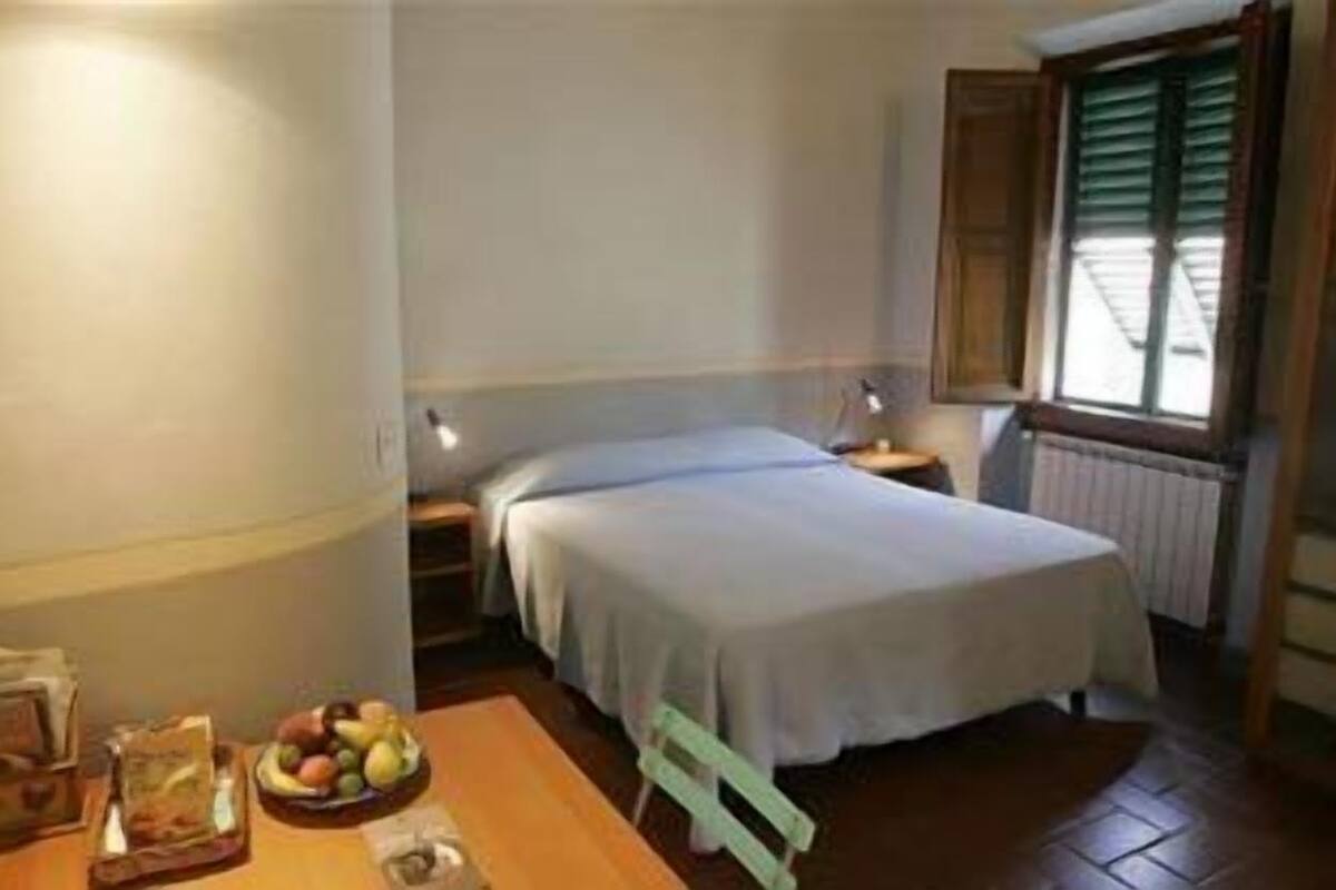 A small, clean room with a single bed, a bedside table with a lamp, a window with shutters, and a fruit bowl on the table.