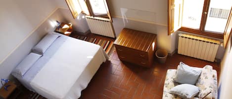 A bedroom with a bed, a small wooden chest, a sofa with cushions, a radiator, and a window with a view of the outside.