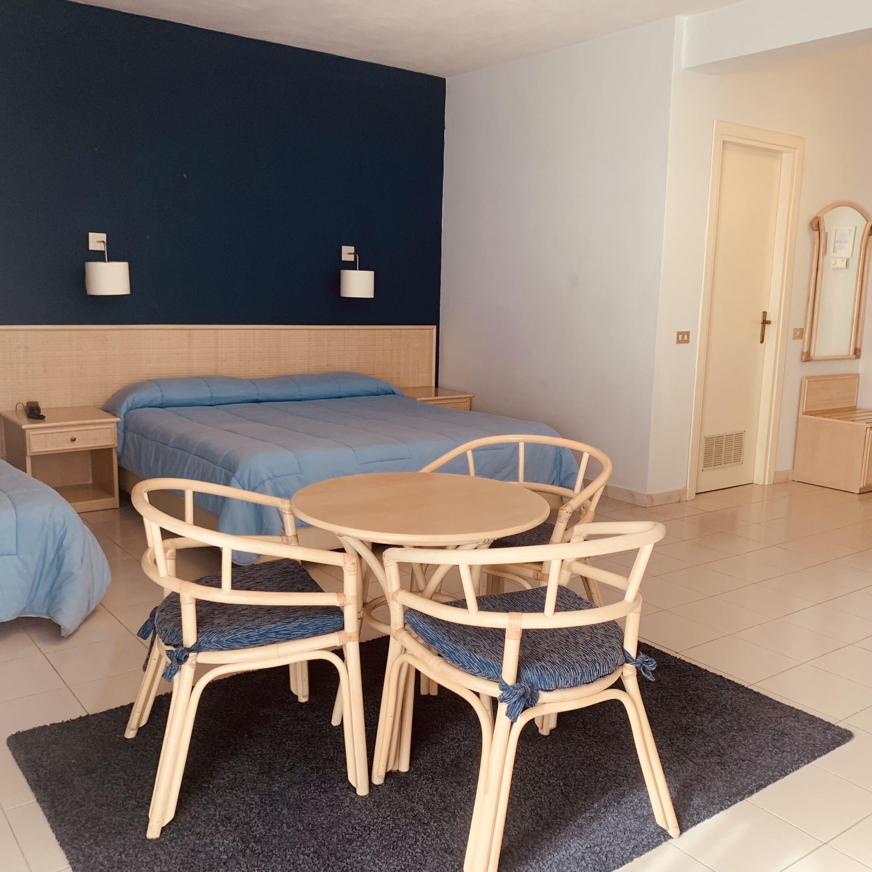 Una habitación de hotel con una cama, dos mesitas de noche, una mesa redonda con sillas, una alfombra oscura y una lámpara de pared.