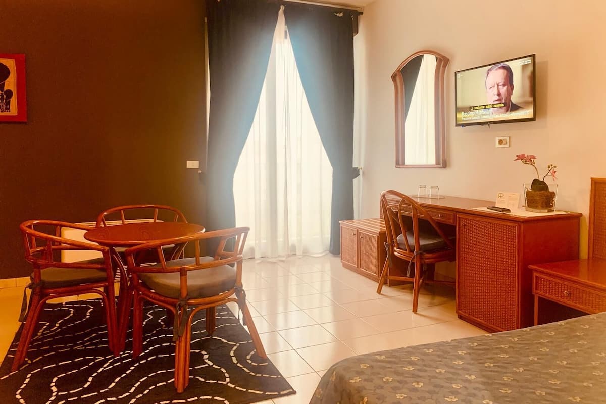 A hotel room with a bed, a desk with a chair, a dining table with chairs, a TV on the wall, and a window with curtains.