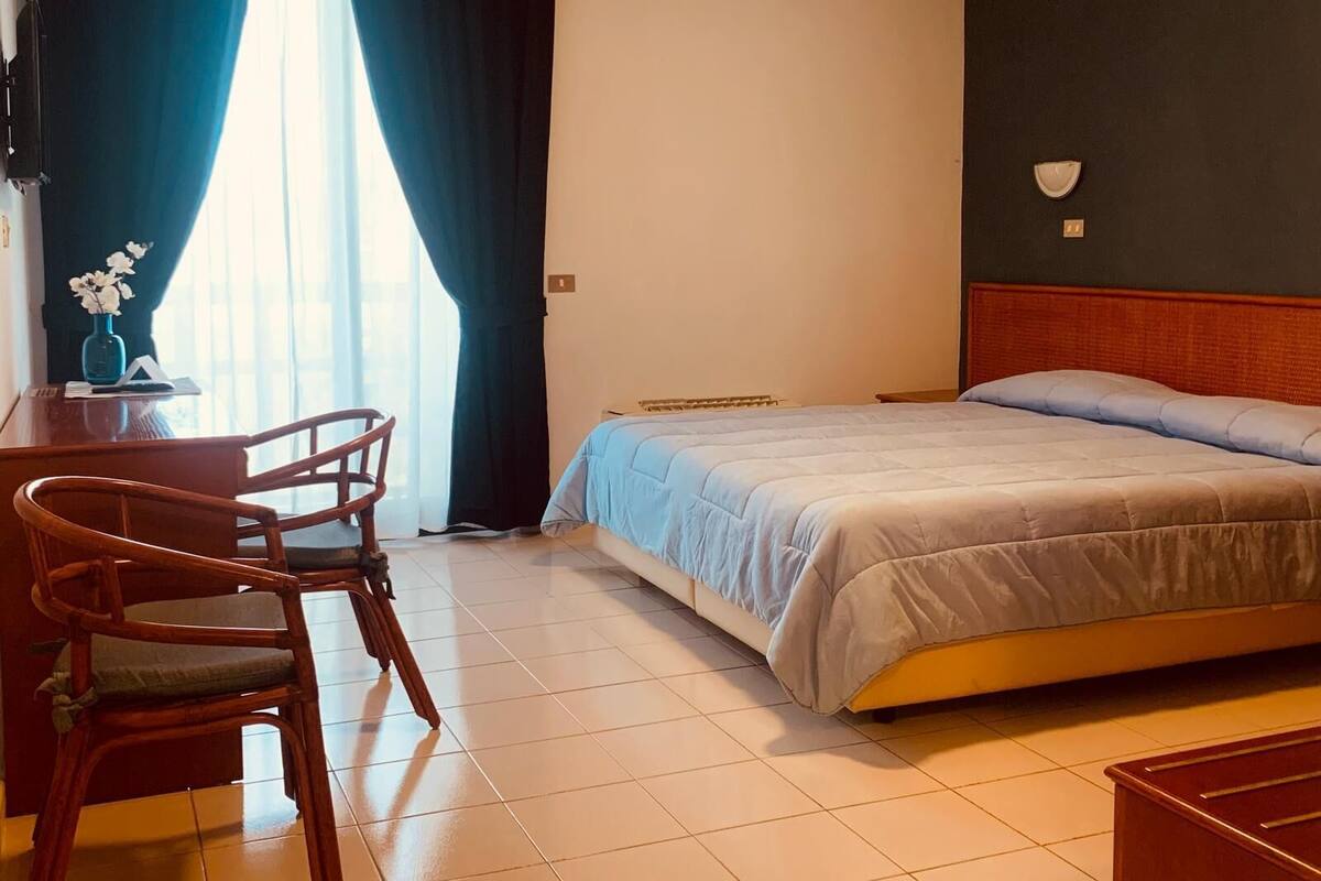 A hotel room with a bed, a desk with a chair, a vase with flowers, and a window with curtains.