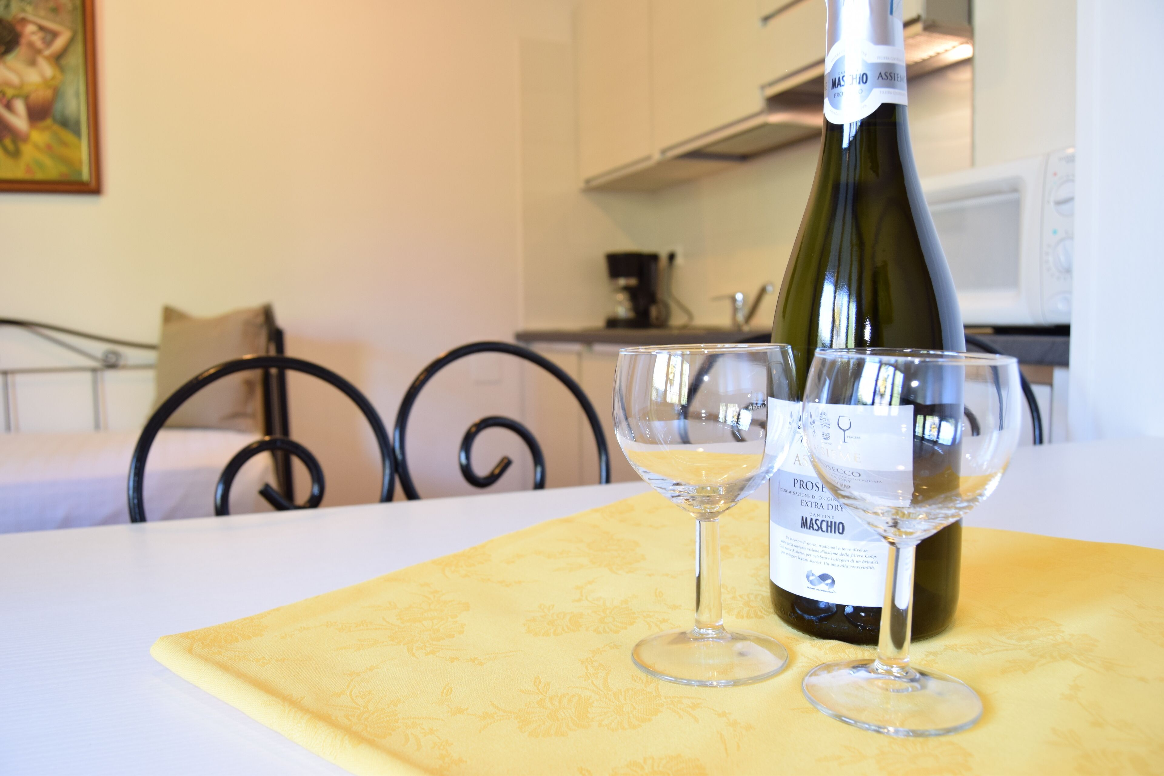 A bottle of Maschio wine and two glasses on a yellow tablecloth in a room with a bed and a chair.