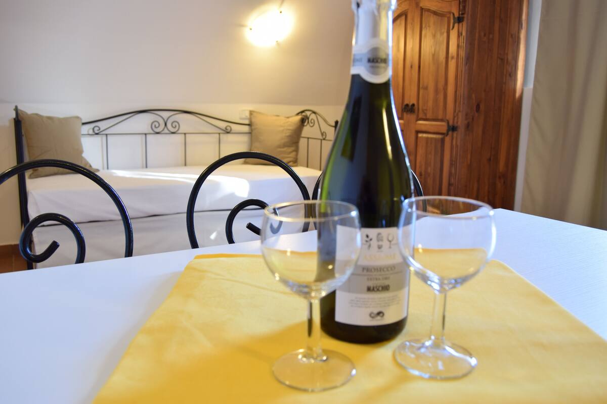 Un lit avec une tête de lit, une porte en bois et une bouteille de vin accompagnée de deux verres sur une table.