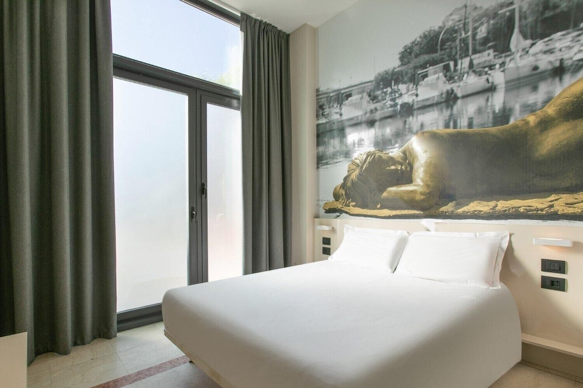 Habitación de hotel con una cama grande, vista al mar a través de una ventana y un cuadro de una figura reclinada en la pared.