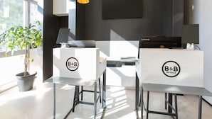 Una moderna zona de recepción de hotel con dos mostradores identificados como "B&B HOTELS", un gran ventanal con vistas a árboles y un televisor de pantalla plana montado en la pared.