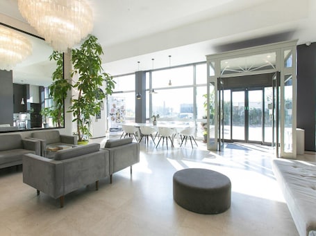 A spacious living area with a modern sofa set, a round ottoman, and a dining table with chairs. There is a large plant and a chandelier.