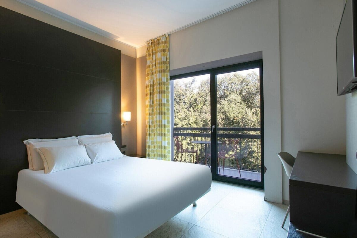 Una habitación de hotel moderna con una cama grande, un balcón con vistas a árboles y un escritorio con una silla.