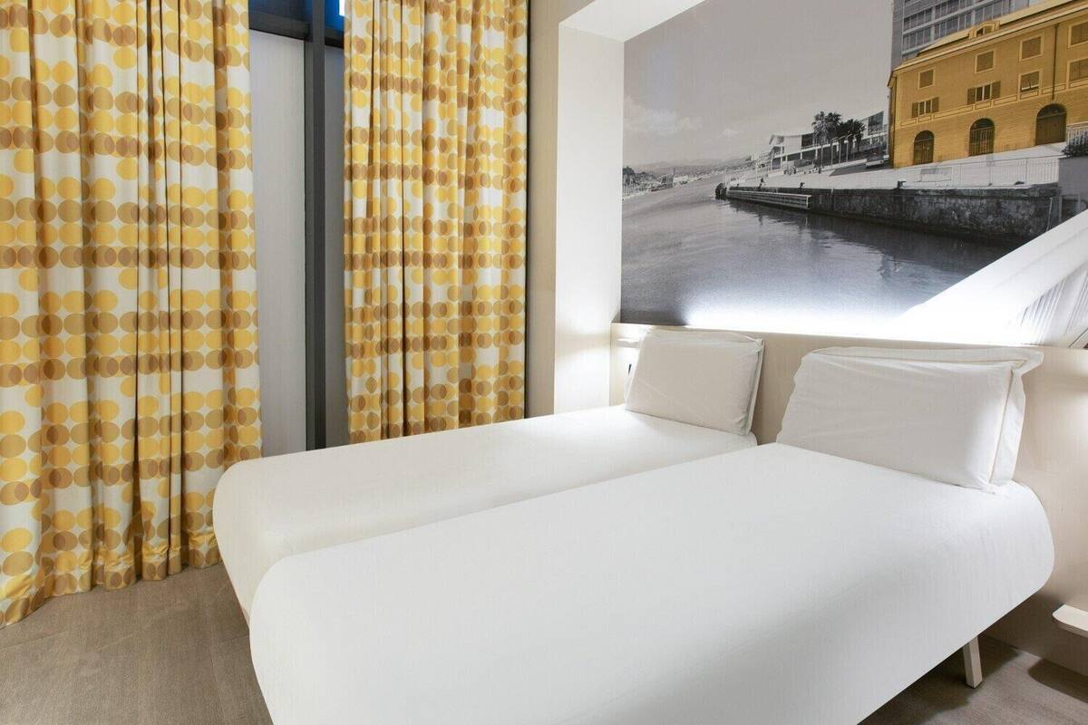 Habitación de hotel con cama, cortinas y un ventanal con vista a la ciudad.