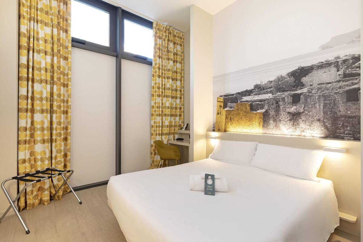 Habitación de hotel con una cama grande, un escritorio y una silla. Hay una ventana con cortinas, un cuadro en la pared y una toalla sobre la cama.