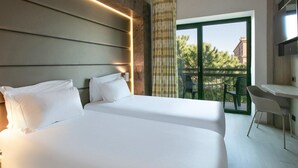 Habitación de hotel con dos camas, un escritorio y un balcón con vistas a árboles y edificios.