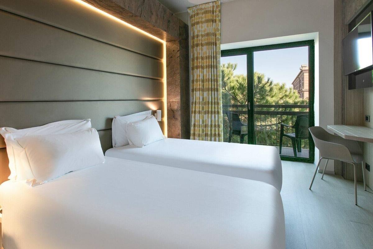 Habitación de hotel con dos camas, un escritorio y un balcón con vistas a árboles y edificios.