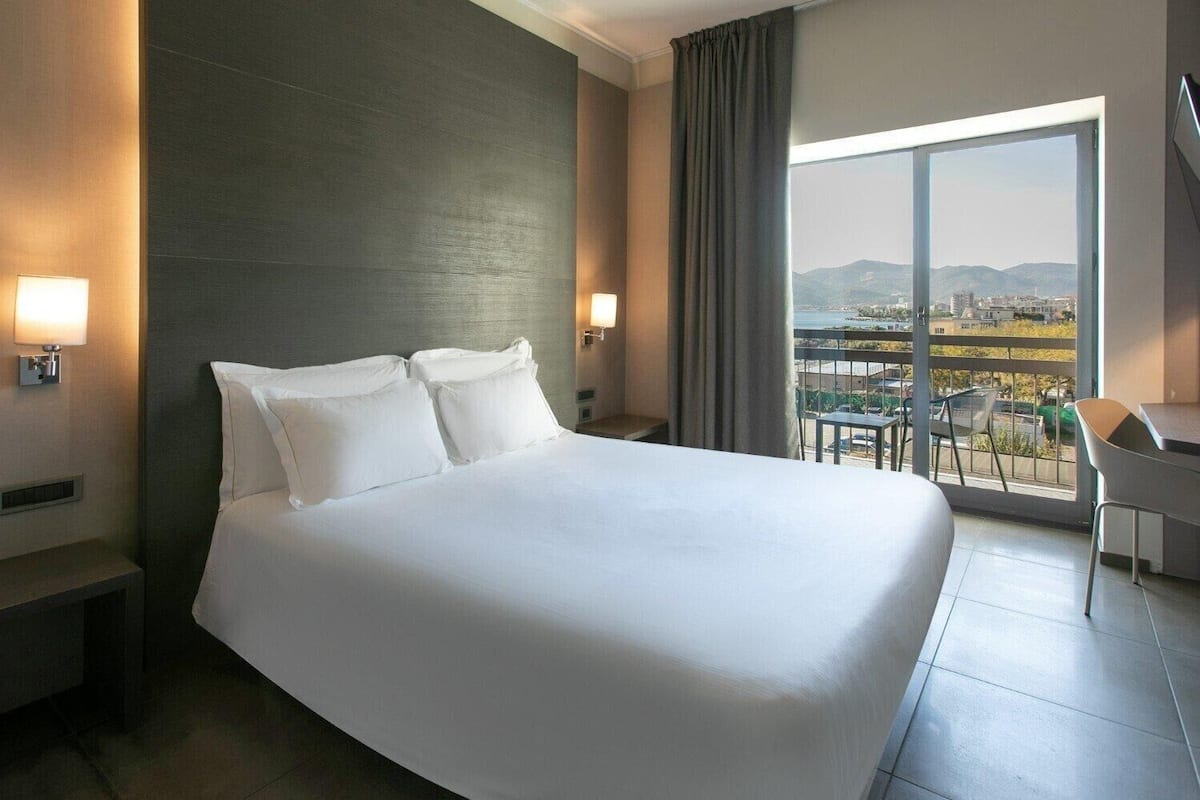 Habitación de hotel con una cama grande, dos lámparas de noche, vistas de la ciudad a través de un ventanal amplio y un balcón con sillas.