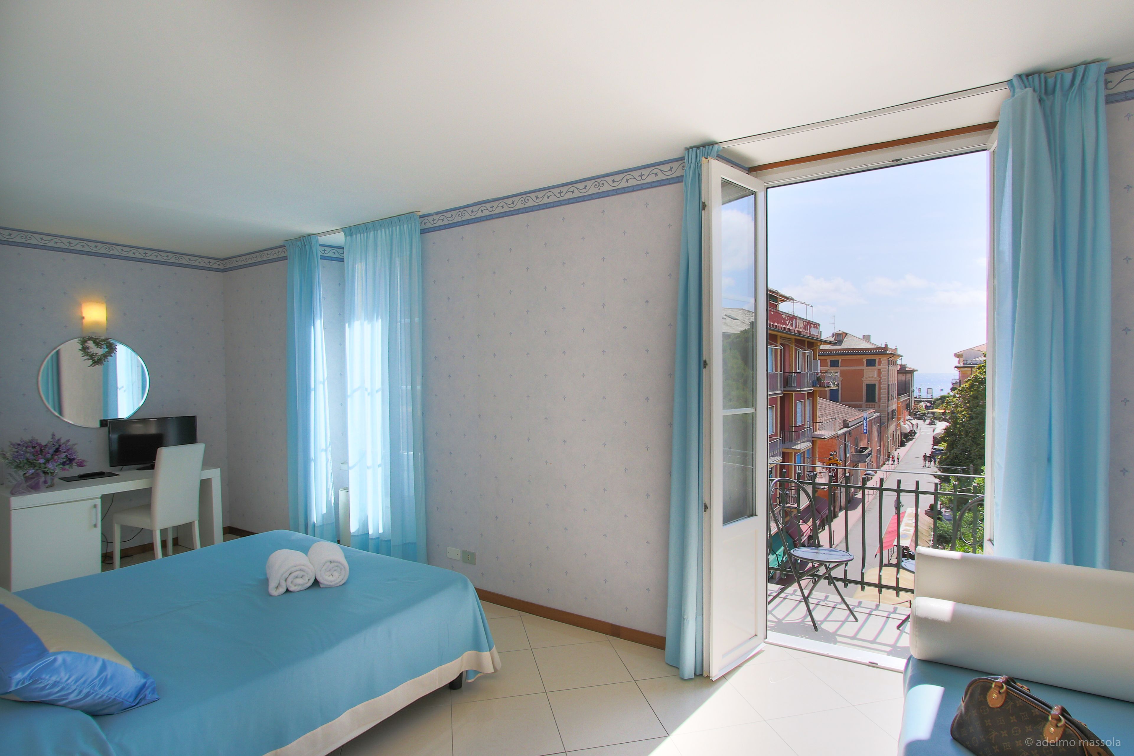 Une chambre à coucher comprenant un lit, un bureau, une chaise, un miroir et un balcon avec vue sur les immeubles.