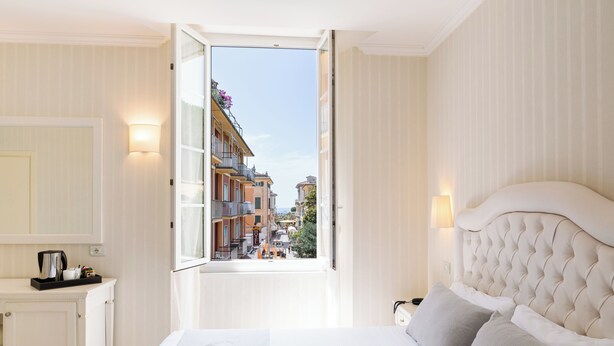 A hotel room with a large bed, a bedside table with a lamp, a view of a cityscape through an open window, and a neatly made bed with towels. - Hotel Nazionale (Levanto)