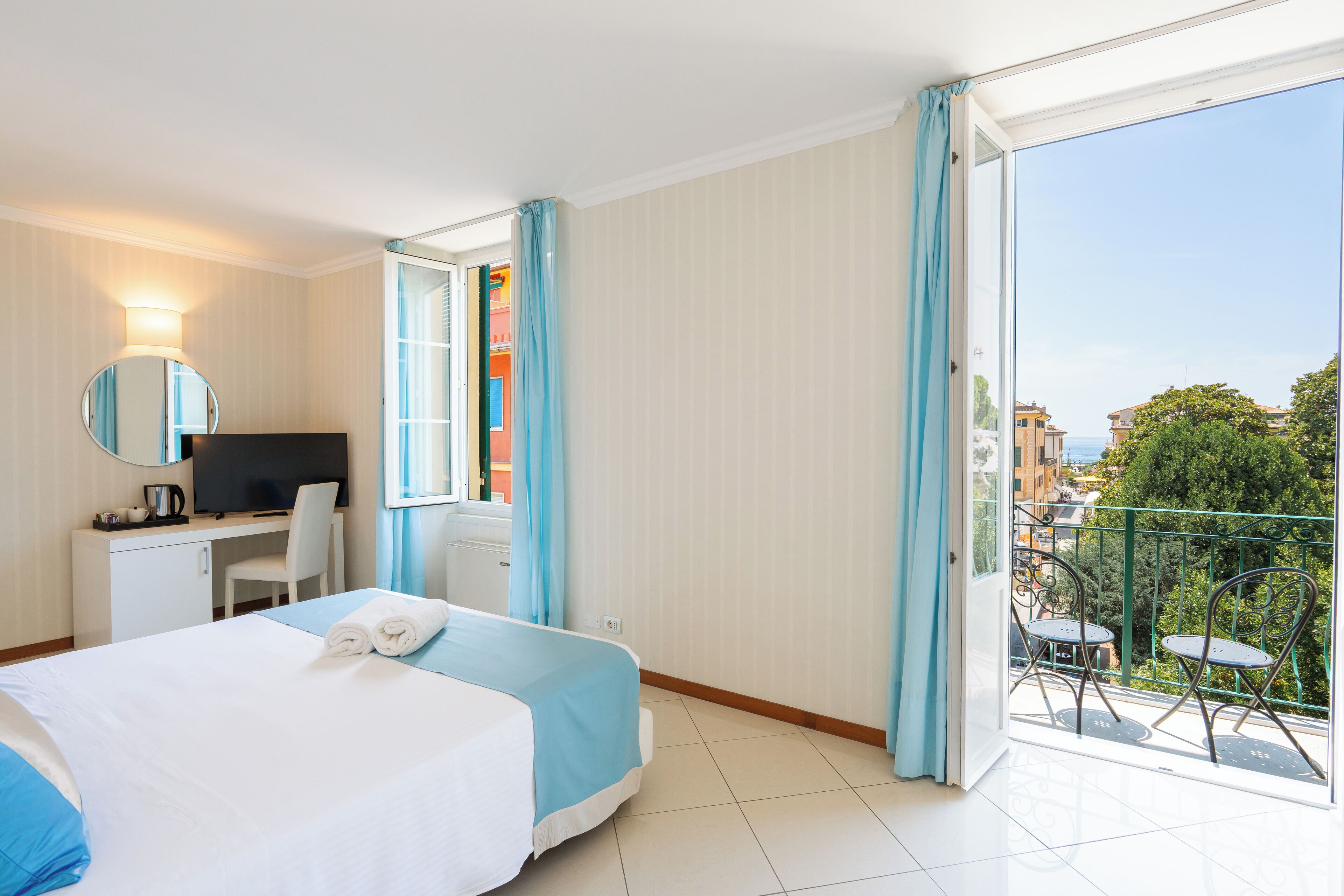 Habitación de hotel con cama, escritorio con televisor, balcón con sillas y vista a la ciudad.