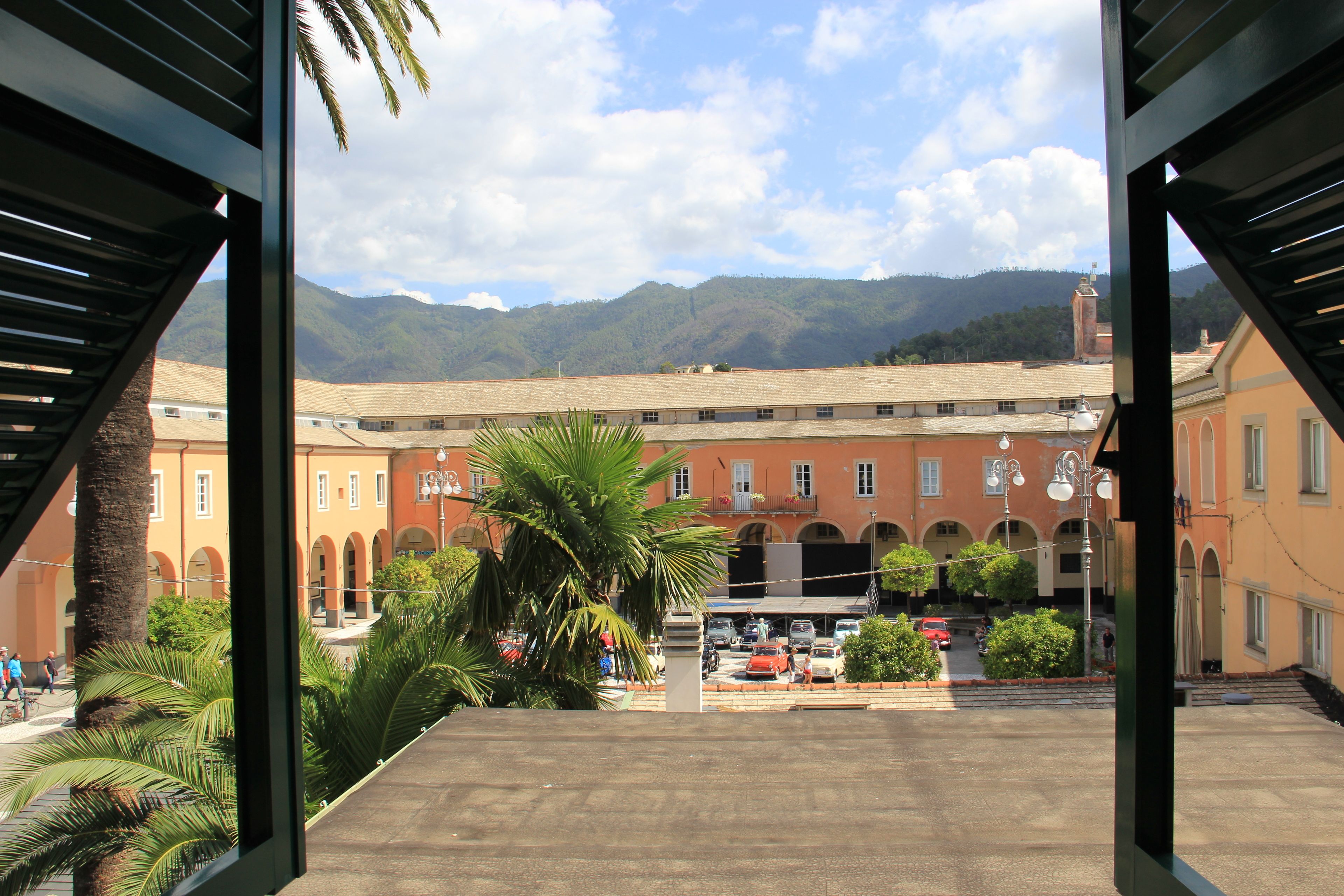 A courtyard with a building featuring arched windows and a red roof, surrounded by palm trees and a clear sky.