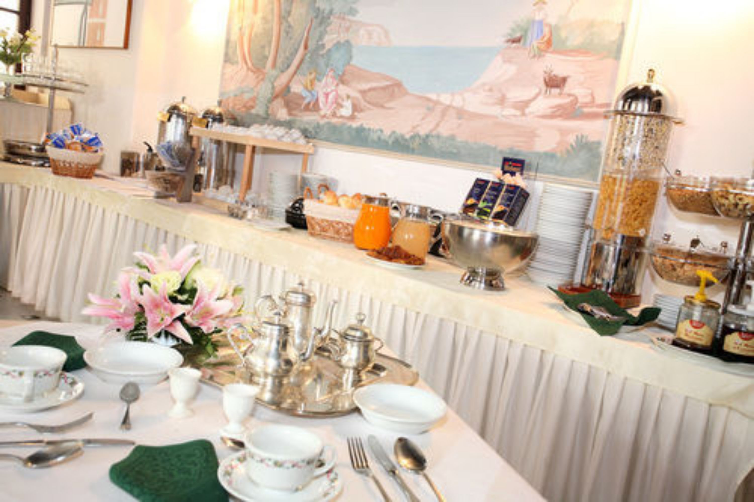 A breakfast buffet setup with various food items and beverages, a decorative painting on the wall, and a neatly arranged table with dining ware.