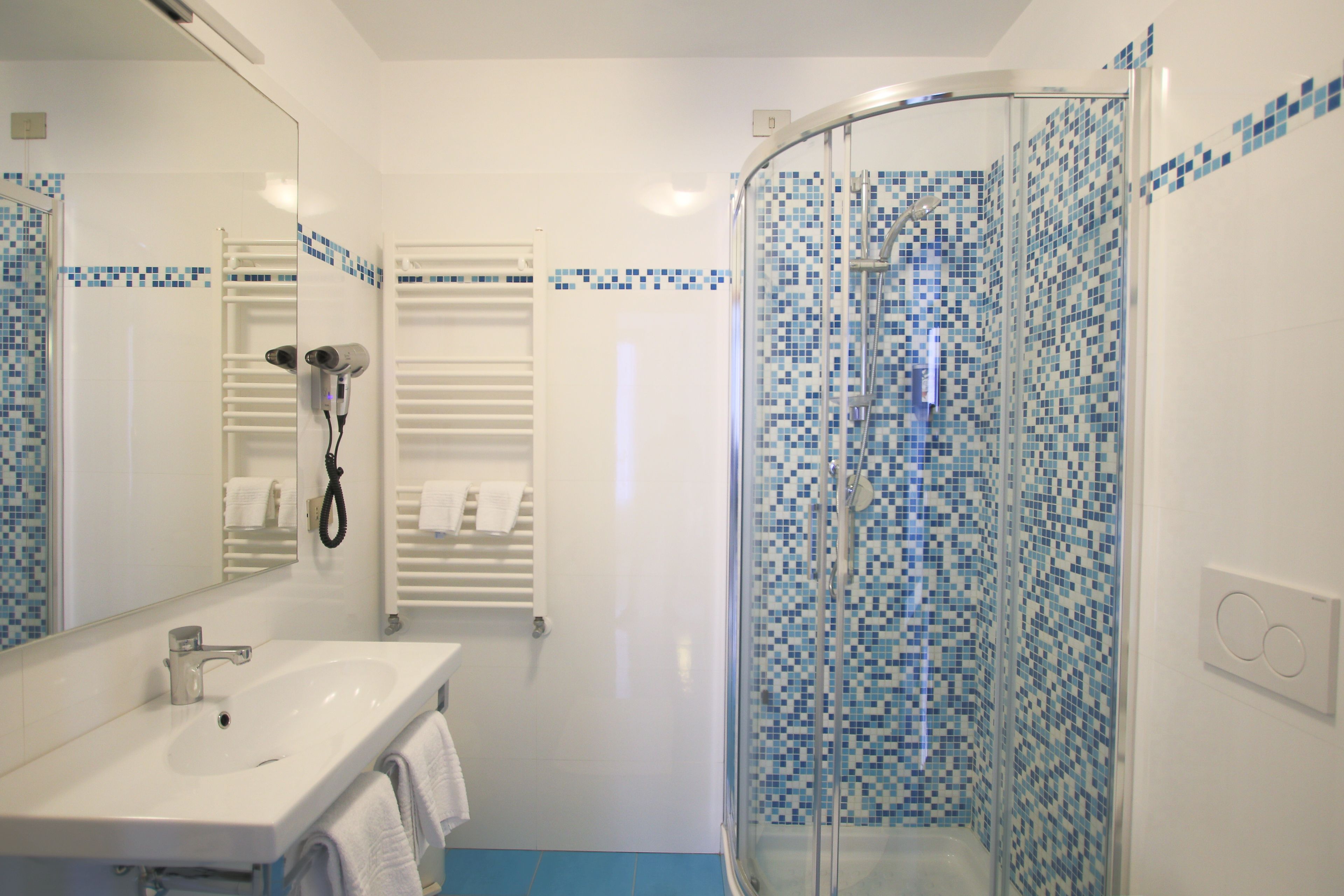 Un baño con lavamanos blanco, un espejo, cerramiento de ducha de vidrio con mosaicos azules y un perchero para toallas.