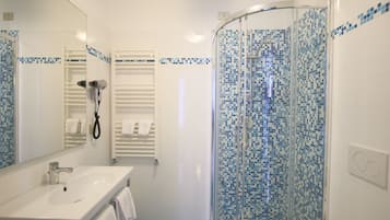 Un baño con lavamanos blanco, un espejo, cerramiento de ducha de vidrio con mosaicos azules y un perchero para toallas.