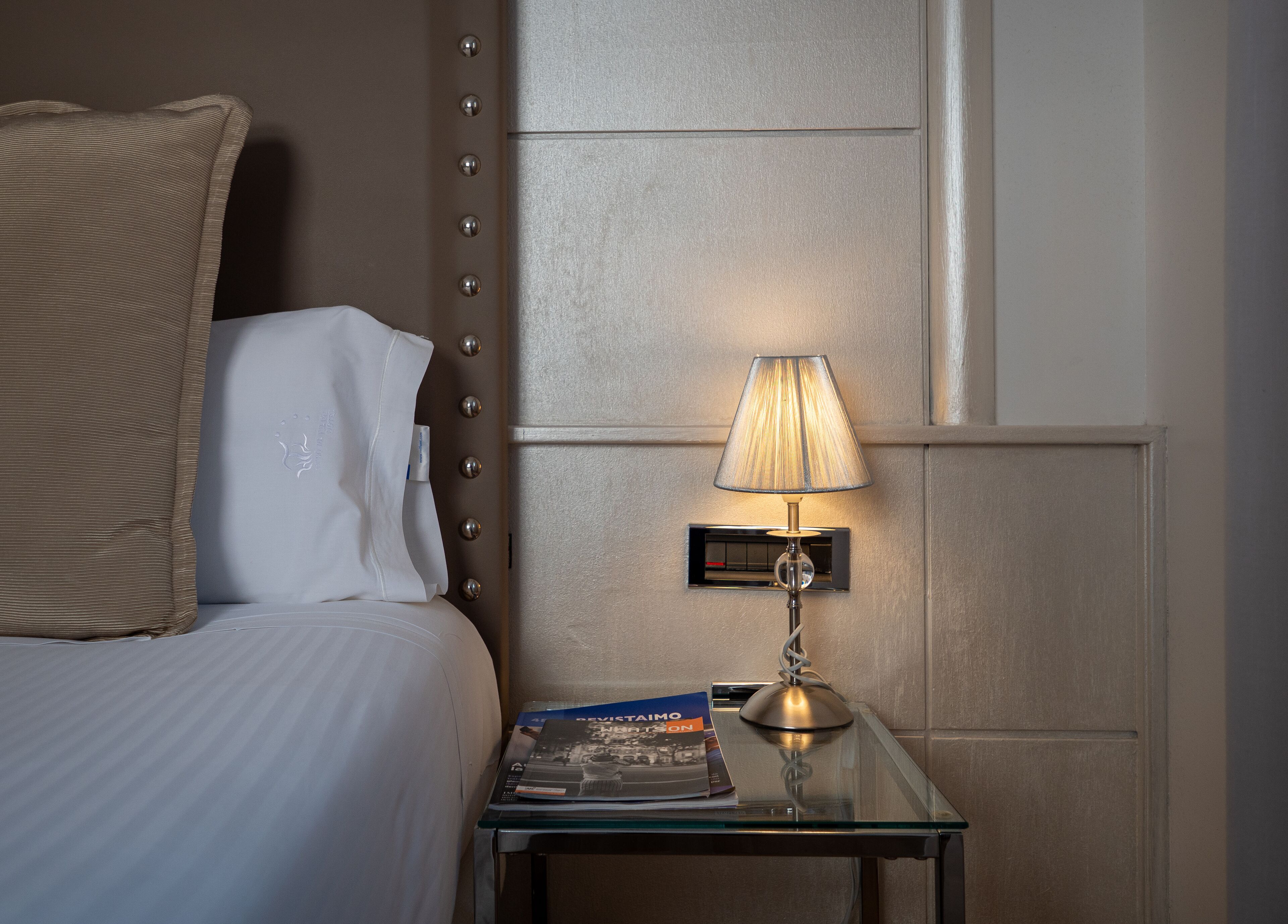 A neatly made bed with a pillow and a bedside table with a lamp and a book.