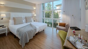 Une chambre d’hôtel avec un grand lit, des tables de chevet, un bureau avec un plateau de collations et une vue sur les arbres à travers de grandes fenêtres.
