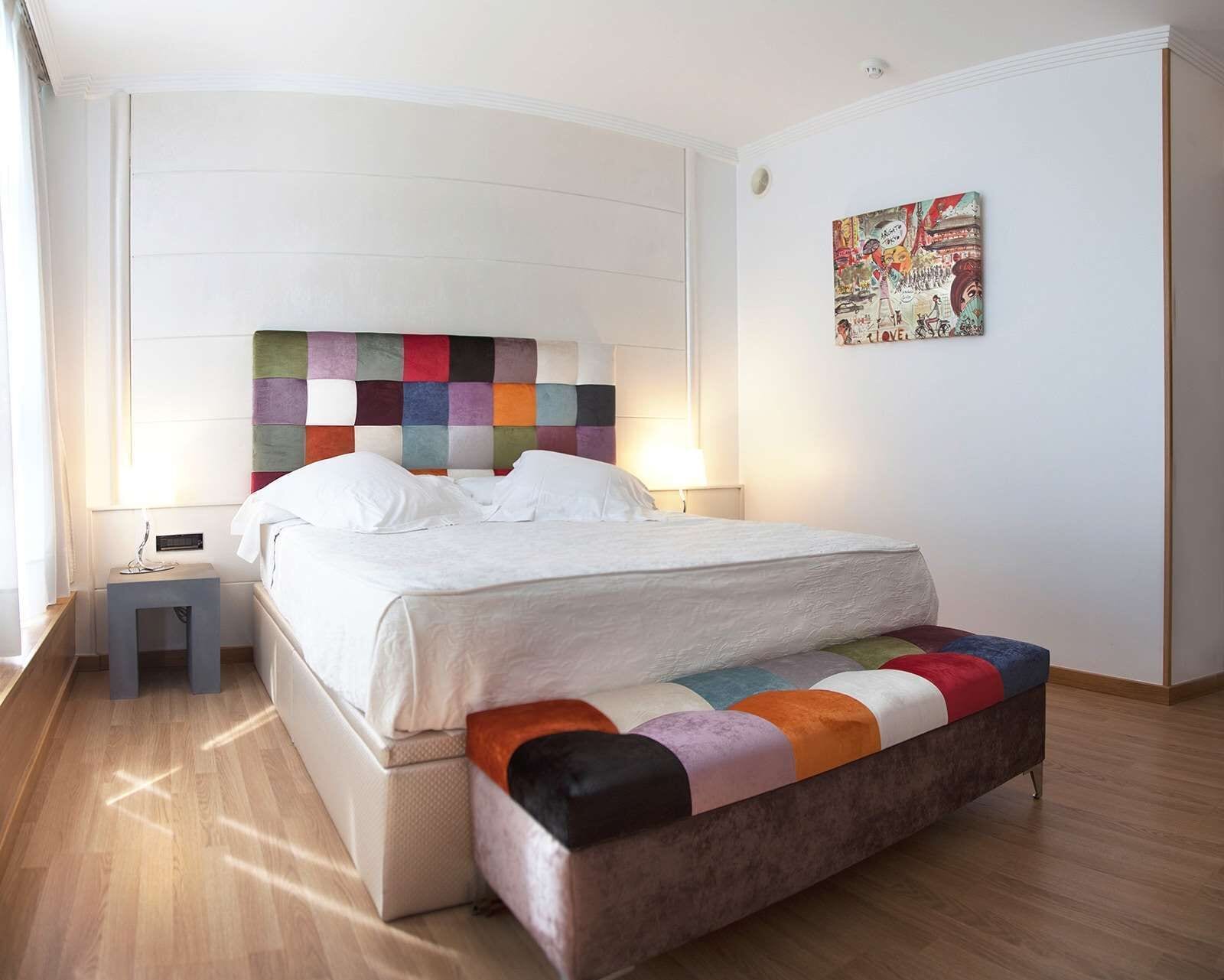 A modern bedroom with a colorful quilted headboard, a white bed, a bedside table with a lamp, and a colorful bench.