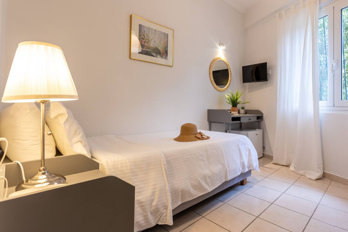 A bedroom with a bed, a bedside table, a lamp, a TV, a mirror, and a window with curtains.