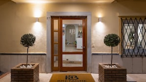 Entrada de hotel con puerta de madera, felpudo con el nombre del hotel y dos macetas a cada lado.
