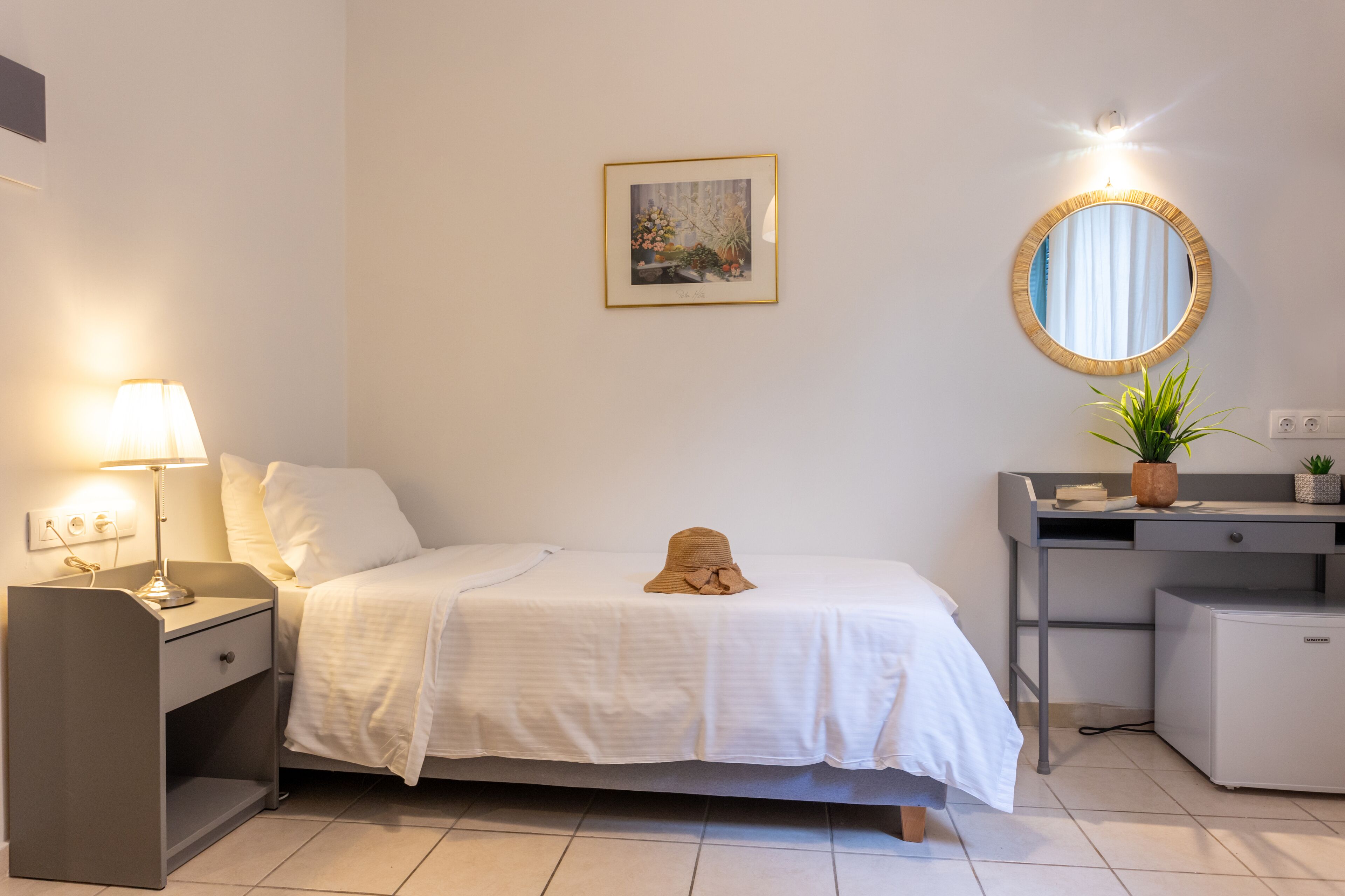 Una camera da letto ordinata con un letto, un comodino, una lampada e una scrivania con uno specchio e piante.
