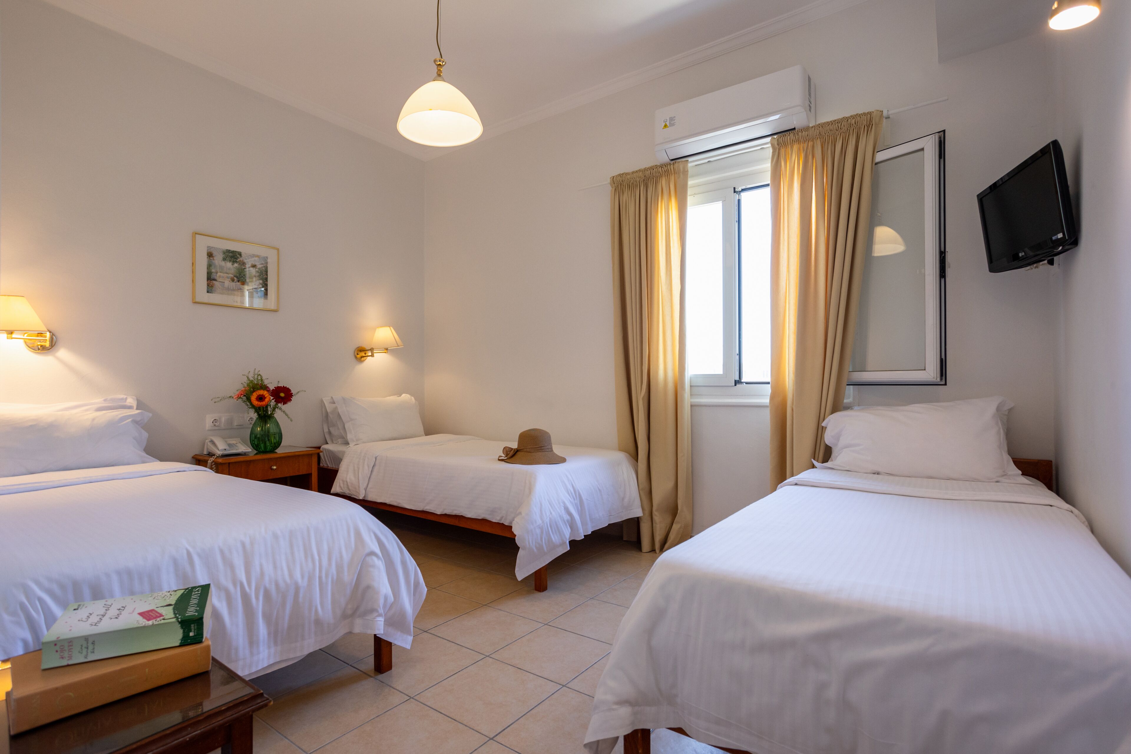 Habitación de hotel con dos camas individuales, una mesita de noche con un libro y un jarrón de flores, un televisor de pantalla plana montado en la pared y una ventana con cortinas.