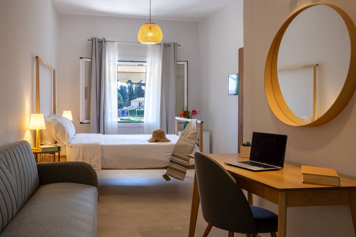 A hotel room with a bed, a desk with a laptop, a chair, a round mirror, and a view of the outdoors.