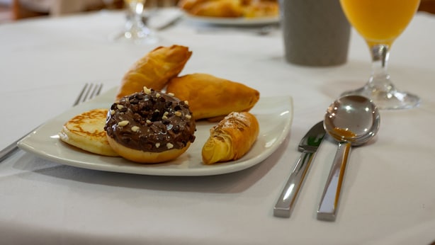 Una escena de desayuno con croissants, un donut cubierto de chocolate y vasos de zumo de naranja sobre una mesa.