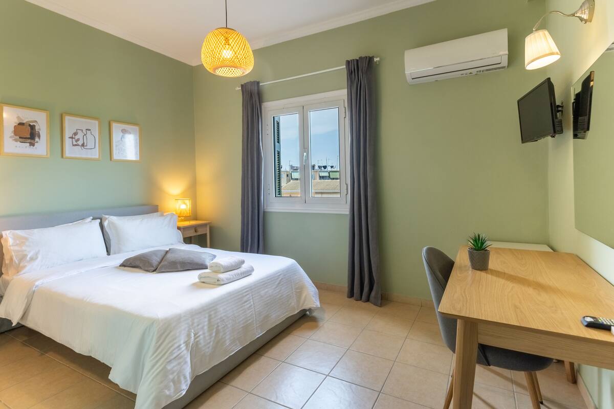 A neatly made bed with white linens and pillows, a bedside lamp, a small table with a plant, and a window with curtains.