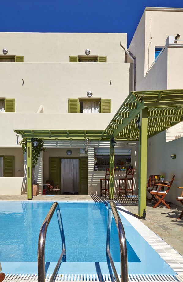 Una piscina con escalera metálica, un pérgola de pilares verdes y un edificio con múltiples ventanas y balcones.