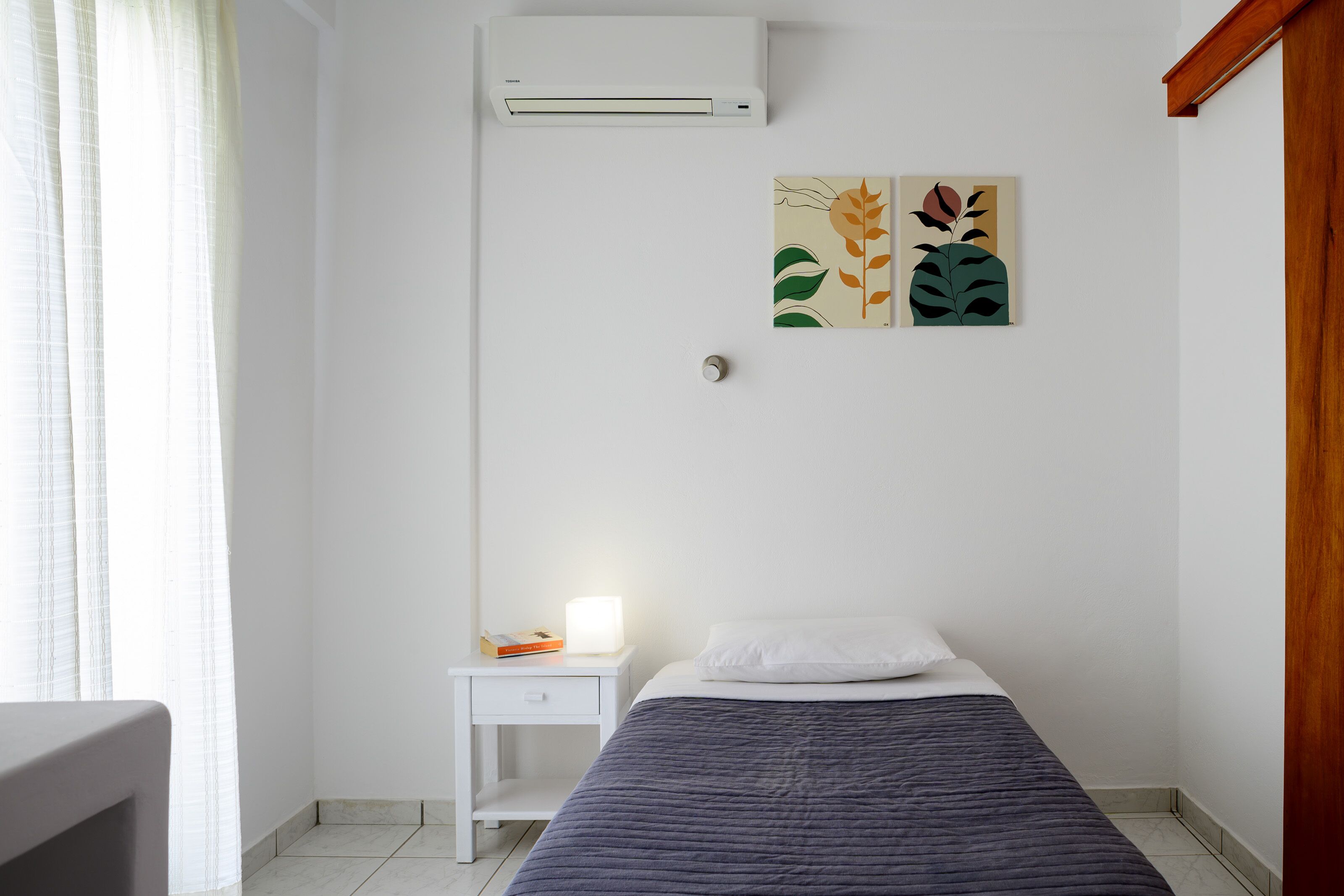 Um quarto com cama, mesa de cabeceira, ar-condicionado fixado na parede e janela com cortinas.