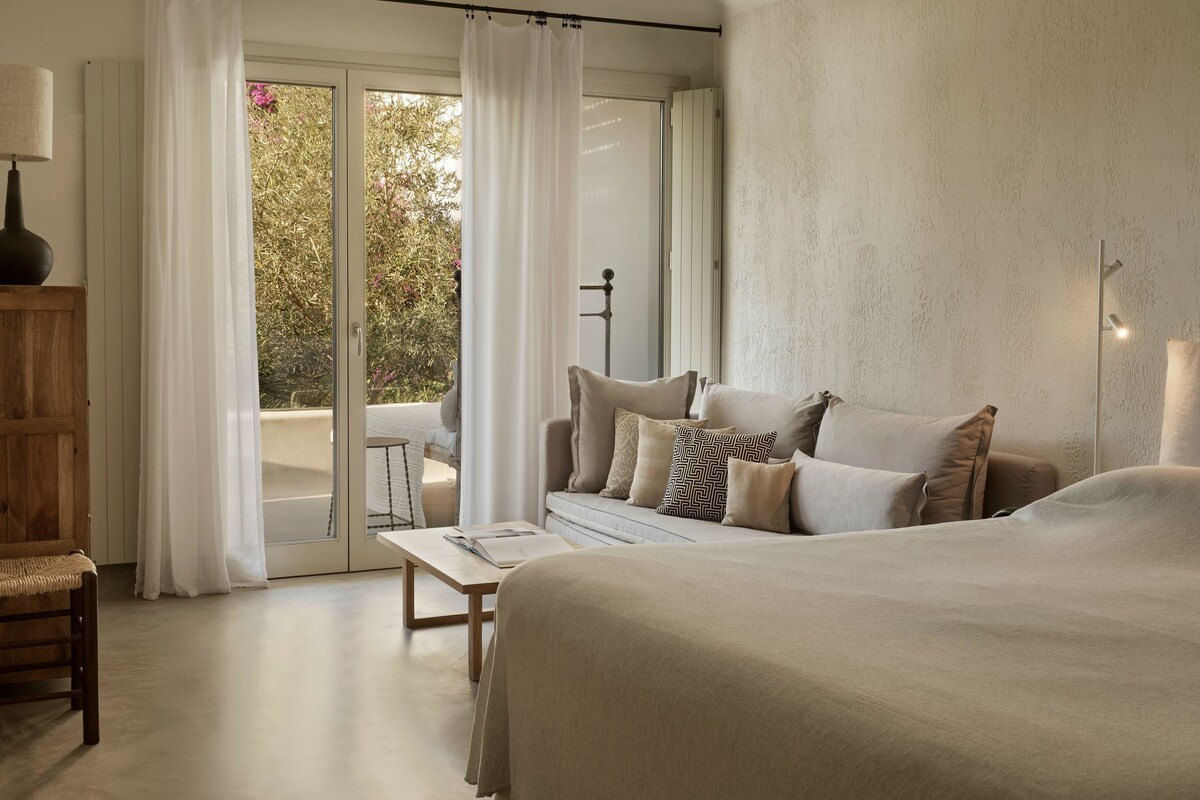 Ein Schlafzimmer mit einem großen Bett, einem Holzstuhl, einem Tisch und Blick auf einen Garten durch eine Schiebetür.